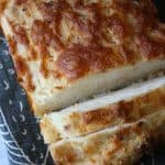 A close-up of a loaf of cheese cider bread on a dark patterned cloth, with two slices cut. The golden, melted cheese is visible on the crust and throughout the bread.