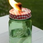 A mason jar citronella candle with a red and white checkered lid sits on a table. A wick burns brightly above the clear liquid inside, while a blurred grassy background sets a summery scene.