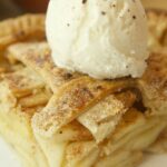 A close-up of a slice of old fashioned apple pie with a lattice crust, topped with vanilla ice cream and a sprinkle of cinnamon on a white plate.