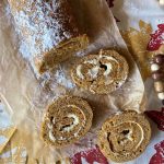 Pumpkin roll recipe with cake mix, dusted with powdered sugar and sliced to reveal a creamy filling, sits on brown parchment atop a fall-themed tablecloth with leaf patterns. Wooden beads accent the cozy autumn display.