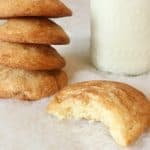 Four stacked snickerdoodle cookies beside a glass of milk, with one cookie in front showing a bite taken out. This cozy scene highlights a snickerdoodle recipe without cream of tartar, set on a soft, white surface.
