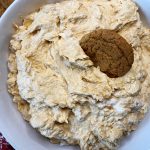 A white bowl filled with pumpkin cream cheese dip, topped with a round brown cookie partially pressed into the creamy mixture.