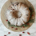 A Cranberry Pound Cake dusted with powdered sugar sits on a wooden board, garnished with fresh rosemary and cranberries, on a white marble surface.