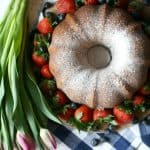 A Mixed Berry Pound Cake dusted with powdered sugar is surrounded by fresh strawberries and blueberries on a checkered cloth, with a bouquet of tulips placed next to the arrangement.