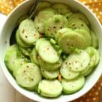 A white bowl filled with Thai cucumber salad, garnished with red pepper flakes and green onions. A spoon rests in the bowl, which sits on a yellow polka dot napkin next to a fork.
