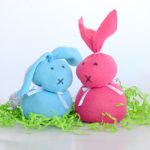 Two homemade sock bunny decorations, one blue and one pink, with drawn faces and ribbon bows, sit on green shredded paper against a white background.