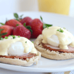Two eggs Benedict on English muffins with ham, poached eggs, and hollandaise sauce, garnished with chives, illustrate How to Make Eggs Benedict—served on a white plate with fresh strawberries in the background.