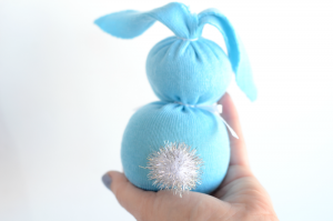 A hand holds a blue, sock bunny-shaped craft with two round sections, floppy ears, and a sparkly silver pom-pom tail against a white background.