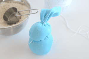 A blue sock bunny made from socks and filled with rice sits beside a metal bowl of rice and a measuring cup. White string ties sections of the bunny, and a ball of white yarn is in the background.