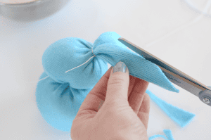 A hand with light blue nail polish uses scissors to cut an ear shape from a blue sock stuffed and tied with string, creating a cute DIY sock bunny craft.