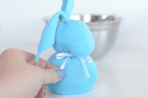 A hand with blue nail polish holds a handmade sock bunny, its soft blue fabric accented by a white ribbon bow around its neck. A metal bowl is blurred in the background.
