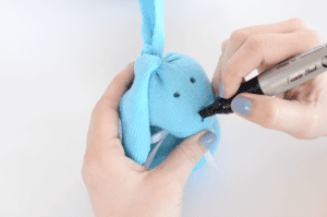 A person uses a black marker to draw eyes on a small blue sock bunny made from fabric, holding it steady with their other hand. The bunny has floppy ears and white pipe cleaner whiskers.