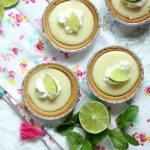 Mini key lime pies topped with whipped cream and lime slices sit on a floral napkin with tassels. Fresh lime halves and mint leaves surround the mini key lime pies on a marble surface, adding a touch of freshness to this delightful scene.