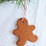 A gingerbread man-shaped applesauce cinnamon ornament with a red and white string hangs beneath green pine branches on white paper decorated with small silver dots.