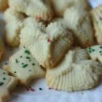 A close-up of holiday cookies, including tree-shaped cookies with green and red sprinkles and golden-brown treats, some made from a classic spritz cookie recipe, all beautifully arranged on a white plate.