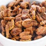 A white bowl filled with Caramelized Pecans, each roasted and coated in a sugar and spice mixture, giving them a glossy, irresistible finish.