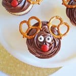 A festive Reindeer Cupcake decorated with chocolate frosting, pretzel antlers, candy eyes, and a red candy nose sits on a white plate atop a gold glittery placemat. Perfect for your holiday Reindeer Cupcakes display!.