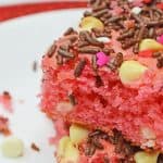 Two pink valentines day cake squares topped with chocolate sprinkles, white chocolate chips, and heart-shaped sprinkles are stacked on a white plate.
