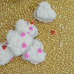 Four heart-shaped cookies with white icing, white sprinkles, and pink and red heart-shaped decorations rest on a textured gold woven surface—perfect alongside your favorite Valentine truffles.