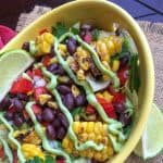 A yellow bowl filled with a vibrant Mexican chopped salad of black beans, grilled corn, red bell peppers, cilantro, and lettuce, topped with green avocado crema and served with lime wedges.