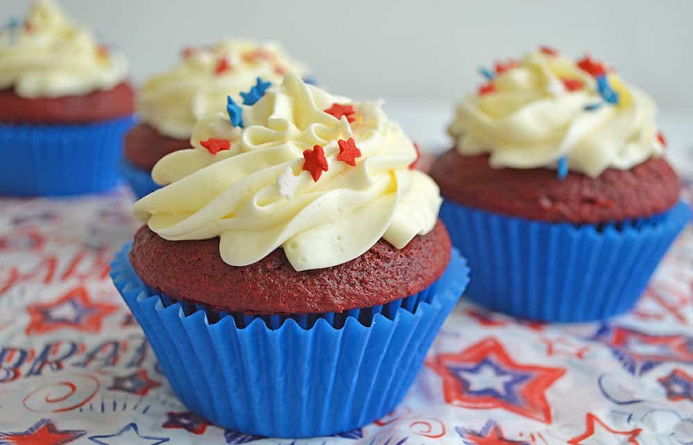 Red velvet red white and blue cupcakes in blue wrappers, topped with swirls of white frosting and star-shaped sprinkles. The background features a festive, star-patterned cloth.