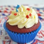 A red velvet cupcake in a blue wrapper topped with swirls of white frosting and festive red, white, and blue star sprinkles—perfect for celebrating with delicious red white and blue cupcakes on a star-patterned napkin.