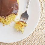 A slice of Boston cream poke cake with chocolate frosting sits on a white plate, with a fork holding a small bite. The plate rests on a decorative woven placemat.