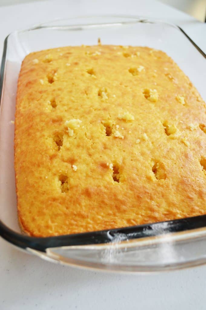 A rectangular pan of golden-brown baked cornbread with evenly spaced holes poked into the surface, reminiscent of a boston cream poke cake, sits invitingly on a white countertop.