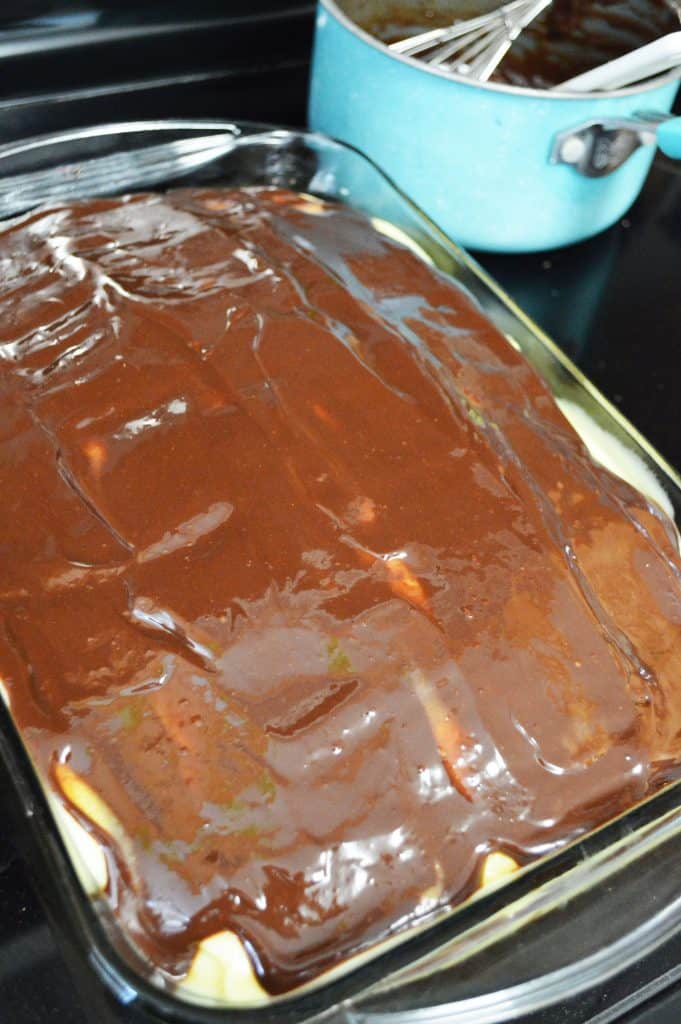 A glass baking dish filled with Boston cream poke cake is covered in a thick layer of chocolate glaze, with a blue saucepan and a whisk visible in the background.