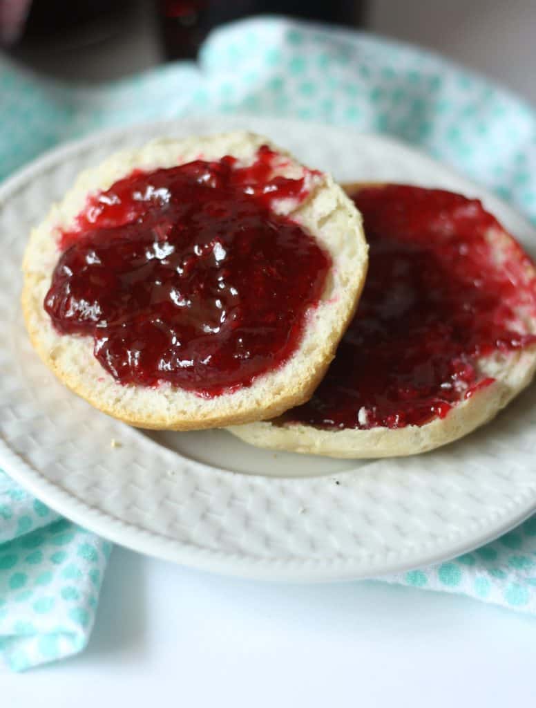A close-up of an English muffin sliced in half, both halves spread with glistening red jelly—perhaps inspired by a classic blackberry jam recipe—on a white plate with a light blue polka dot cloth in the background.