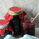 Three jars of dark red jam, made from a classic blackberry jam recipe—two covered with red and white polka dot cloths, one open with a gold spoon. A blue and white towel sits in the foreground on a marble surface.