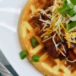 close up of a plate with a cornbread waffle covered in chili, cheese, sour cream, and green onions.