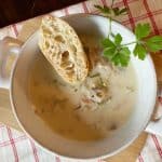 White bowl of clam chowder with bread and parsley garnish on a red plaid towel.