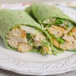 Close-up of two spinach wraps packed with chicken, lettuce, croutons, and shredded cheese on a white plate.