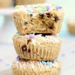 Three chocolate chip cookie cupcakes are stacked on top of each other, each topped with white icing drizzle and colorful pastel sprinkles. The top cupcake has a bite taken out, revealing its soft, moist texture inside.