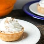 A mini pumpkin pie topped with swirled whipped cream and a sprinkle of cinnamon sits on a white plate, with another mini pumpkin pie and a pumpkin in the background.