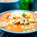 A close-up of a blue bowl filled with traditional minestrone soup recipe, topped with grated cheese and fresh herbs. The colorful soup features beans, greens, and chopped vegetables. A spoon and another bowl are blurred in the background.