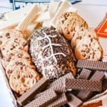 A football-shaped peanut butter cheesecake dip covered in chocolate chips with white icing laces, surrounded by chocolate chip cookies and wafer cookies on a tray.