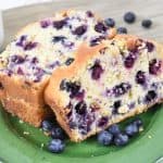Two thick slices of blueberry loaf cake, made from a delicious blueberry bread recipe, sit on a green plate with fresh blueberries scattered around. The golden brown cake is packed with juicy blueberries.