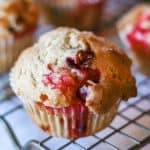 A close-up of a golden-brown Panera Cranberry Orange Muffin with visible bits of red fruit sits on a metal cooling rack, with more muffins blurred in the background.