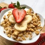 A bowl of instant pot yogurt topped with granola, banana slices, and a halved strawberry, with two whole strawberries in the background and a red cloth napkin on the side.