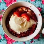 A round white dish filled with Strawberry Shortcake Dump Cake, topped with a scoop of vanilla ice cream and sliced strawberries, sits on a blue tablecloth patterned with red and pink roses.