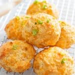 Six golden, cheesy biscuits—using the iconic Red Lobster Biscuit Recipe—are topped with chopped green onions and arranged on a cooling rack, with a blurred kitchen utensil in the background.