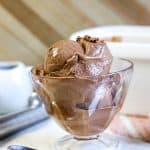 A glass dessert cup filled with two scoops of creamy no churn chocolate ice cream sits on a table, with a metal scooper, cherries, and a white container in the background.