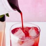 A close-up of a glass filled with ice while a red liquid, possibly blackberry simple syrup, is being poured into it. Other beverages in glasses are visible nearby against a pink and white background.