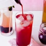 A glass filled with ice is being poured with a deep red juice, likely mixed with blackberry simple syrup, with another glass of pink beverage and jars nearby, all set on a light pink napkin against a pink background.