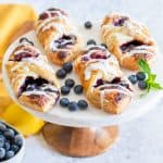 Blueberry pastries on a cake stand with a bowl of blueberries and a lemon nearby.