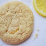 A soft, round chewy lemon sugar cookie with a slightly cracked surface sits on a white napkin next to a lemon slice and a few cookie crumbs.