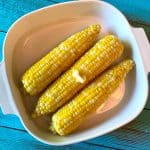 Three ears of cooked corn on the cob with melting butter are placed in a white dish on a bright blue wooden surface, perfect for serving after learning how to cook corn on the cob in the microwave.
