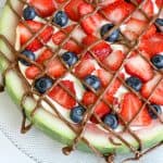 A refreshing watermelon pizza: a round watermelon slice topped with cream, fresh strawberries, blueberries, and a drizzle of chocolate, served on a textured glass plate.